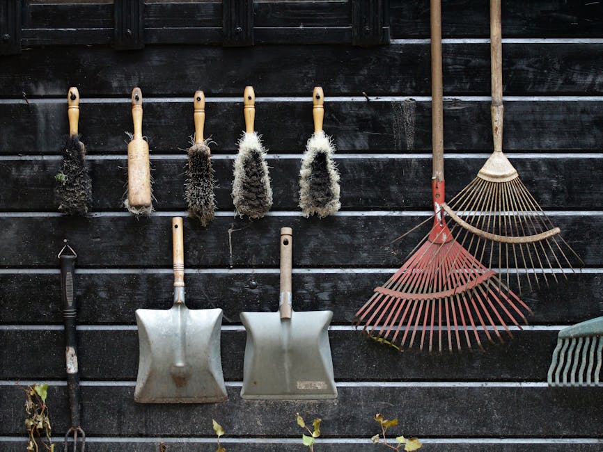 A wooden and metal garden trowel and a pair of pruning shears rest inside a green wire basket with wooden handles, placed on a stone garden wall. Surrounding the basket are various plants, including s